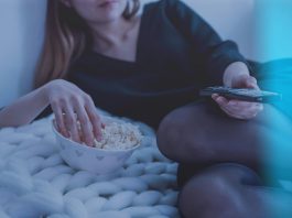 Woman in white bed holding remote control while eating popcorn