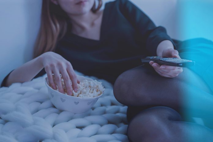 Woman in white bed holding remote control while eating popcorn